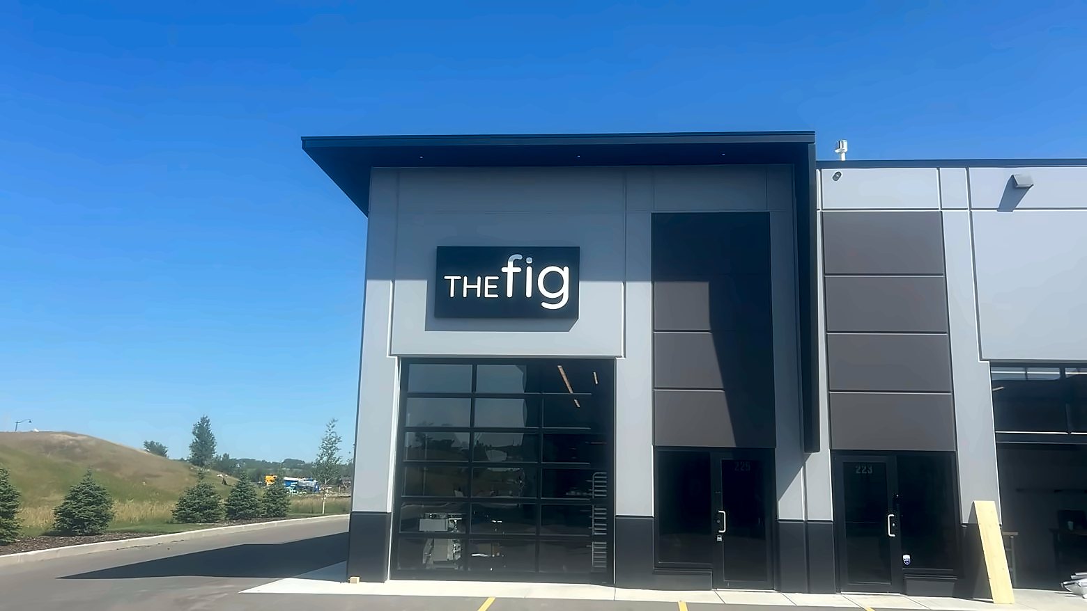 The Fig at the Foundry: a grey and charcoal storefront with a lit sign above a glass overhead door, under a clear blue sky